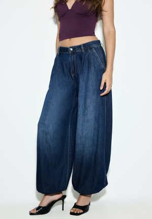 Femme portant un jean en denim bleu foncé à jambes larges, des chaussures à talons hauts ouvertes aux orteils noires, et un top court violet.