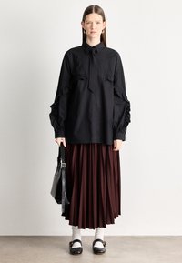 Črna srajca s gumbi in nabranimi rokavi, kombinirana s plisirano bordo krilo. Črna torbica in bele nogavice dopolnjujejo obleko.