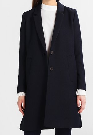 Woman wearing a dark navy long coat with two buttons over a white top with ribbed cuffs, standing against a plain background.