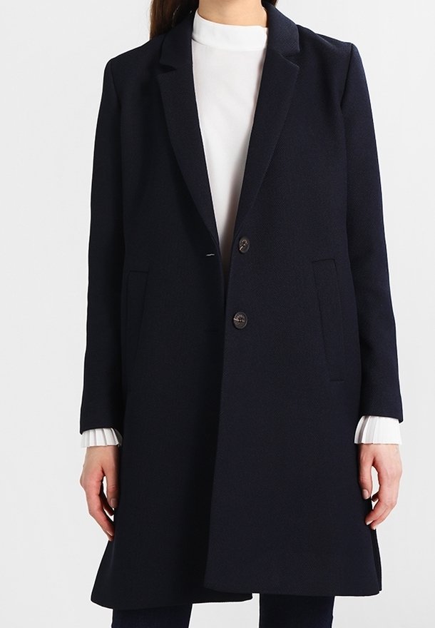 Femme portant un long manteau bleu marine foncé à deux boutons sur un haut blanc à poignets côtelés, debout devant un fond uni.