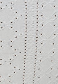 White textured fabric with diamond and dot patterns, featuring small brown accents. The surface is smooth with intricate details.