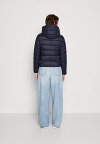 Person med en marinblå pufferjacka med huva och ljusblå vida jeans, som står vänd mot en enkel vit vägg.