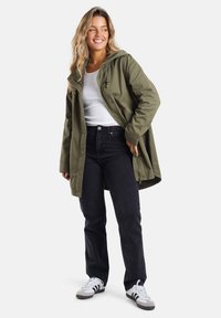 Olivgrüne Kapuzenjacke mit einem Reißverschluss und Knopfverschluss, kombiniert mit schwarzen Jeans und weißen Sneakers. Die Jacke hat einen verstellbaren Kordelzug.