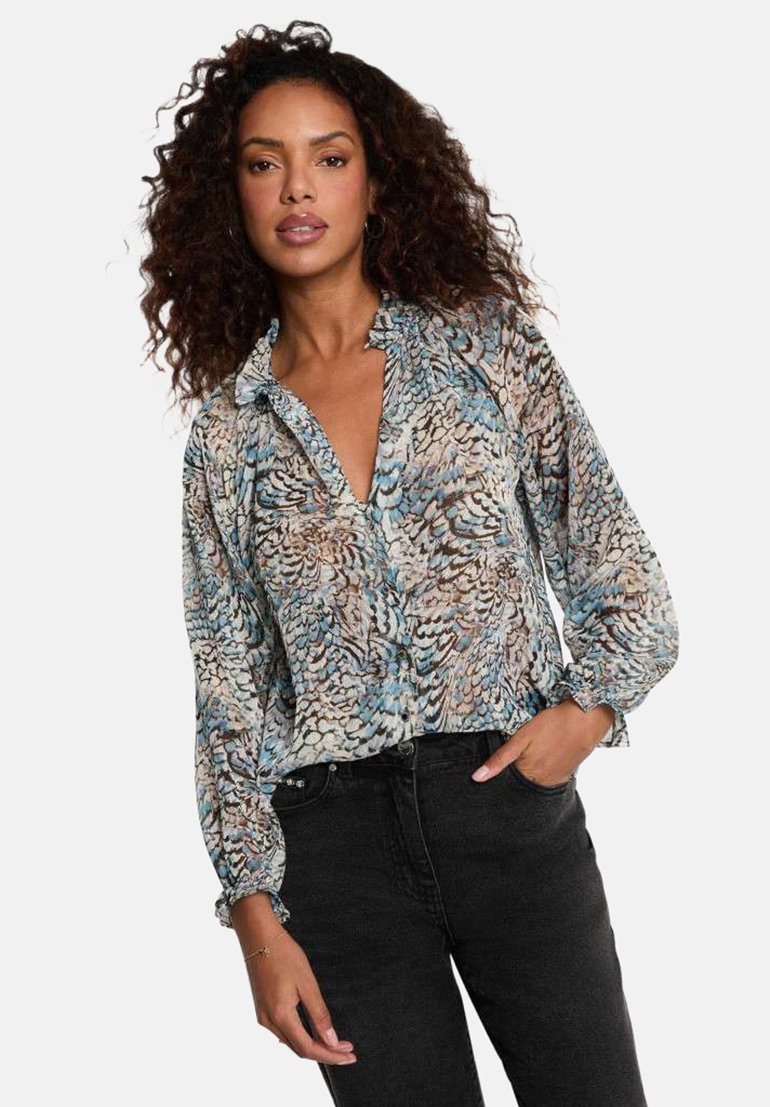 Donna con capelli ricci che indossa una camicetta a maniche lunghe con motivo blu, marrone e beige e jeans neri, in piedi con una mano in tasca.