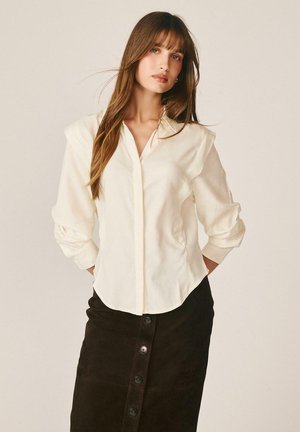 Woman with long brown hair wearing a white buttoned blouse and a black buttoned skirt standing with hands behind her back.