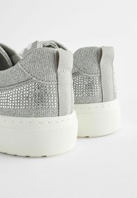 Grå sneakers med en texturerad ovandel prydd med silverfärgade rhinestones, platta vita gummisulor och tyghälar för enkel påtagning.