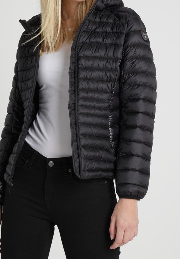 Frau trägt eine schwarze gesteppte Daunenjacke über einem weißen Shirt und schwarzen Hosen und steht vor einem schlichten Hintergrund.