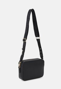 Czarna skórzana torebka na ramię o prostokątnym kształcie, z suwakiem i regulowanym paskiem z nadrukiem logo oraz złotymi akcentami.