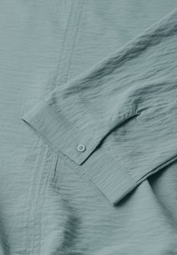 Camicia azzurra a maniche lunghe con polsini abbottonati, morbida al tatto e con un sottile motivo stropicciato e dettagli di cucitura visibili.