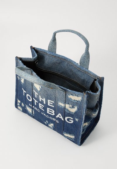 Borsa tote in denim blu con toppe invecchiate, dotata di un paio di manici e una tasca interna con zip nera. Logo stampato in bianco.