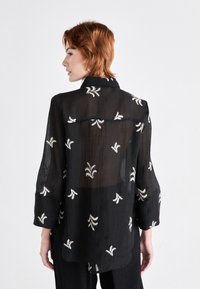 Bluză neagră din material transparent, cu broderie florală albă, mâneci lungi, guler și o croială lejeră, având un tiv ușor curbat la spate.