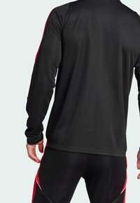Hombre vestido con una camiseta atlética de manga larga negra y pantalones negros con rayas rojas, visto desde atrás sobre un fondo claro.