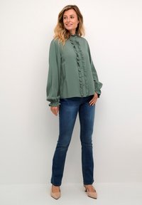 Light green blouse with ruffle detailing, high neckline, and long sleeves, paired with blue jeans and nude pointed heels. Soft, flowing fabric.