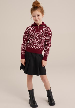 Maglione bordeaux con motivi ondulati bianchi, design con zip al collo. La gonna nera plissettata e gli stivali neri alla caviglia completano l'outfit.