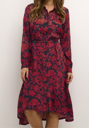 Femme portant une robe bleu marine à imprimé floral rouge, manches longues, boutonnée devant et ceinture nouée à la taille.