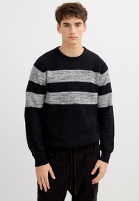 Suéter negro de tejido texturizado, con rayas horizontales grises. Cuello redondo y mangas largas, combinado con pantalones oscuros.