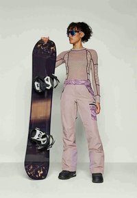 Persoon die een zonnebril en winteruitrusting draagt, terwijl hij een zwarte snowboard met bindingen vasthoudt, staand tegen een effen lichte achtergrond.