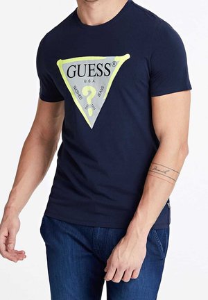 Homme portant un t-shirt bleu marine avec un grand logo triangulaire Guess et un jean bleu, montrant un tatouage sur l'avant-bras et une pose décontractée.