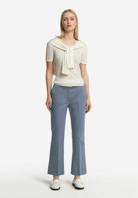 Vit kortärmad tröja med knutkrage, tillsammans med ljusblå vida byxor. Modellen har på sig vita loafers. Enkel, strukturerad design.