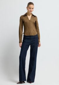 Woman wearing a fitted brown long-sleeve blouse with front buttons and dark wide-leg jeans, standing against a plain light background.