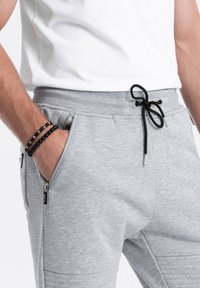 Gray sweatpants made of soft fabric with a drawstring waistband, side zip pockets, and textured knee accents. Black bead bracelet on wrist.