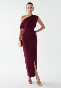 Bordový šaty na jedno plece s drapovaným živôtom, priliehavým pásom a bočným rozparkom. Skombinované so zlatým náramkom a textúrovanou kabelkou, nosené s remienkovými podpätkami.