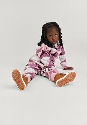 Rosa-weißer Camouflage-Jumpsuit mit Reißverschluss, kombiniert mit braunen Stiefeln mit strukturierten Sohlen und rotem Branding.