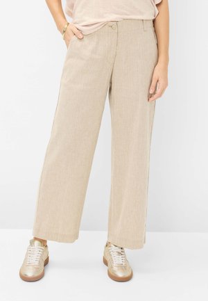 Kvinde iført beige bukser med vide ben og subtile nålestriber, lyserød top, gyldt armbånd og metalliske guld sneakers på hvid baggrund.