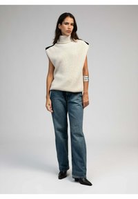 Un top sans manches en tricot couleur ivoire avec des accents noirs aux épaules, associé à un jean en denim bleu ample et des bottes noires à bouts pointus.