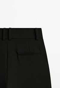 Gros plan sur un pantalon noir sur mesure montrant la ceinture, les passants de ceinture et une poche arrière passepoilée sur fond blanc.