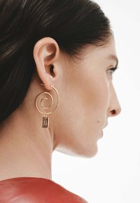 Pendiente de aro en espiral dorado con un charm de candado rectangular colgante. Textura suave, diseño minimalista. Modelo vistiendo un top de cuero rojo.