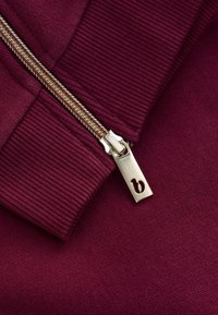 Primo piano di una cerniera dorata su un capo di tessuto a coste di colore rosso scuro, cursore della cerniera con una piccola incisione di lettera minuscola "b".