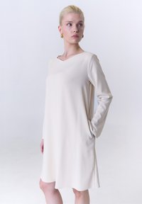 Langärmliges V-Ausschnitt-Kleid aus weichem beigem Stoff, mit seitlichen Taschen, lockerer Passform und knielanger Säume. Glatte Textur.