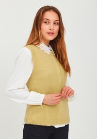 Knit sleeveless sweater in light green, featuring a ribbed hem and round neckline, layered over a white blouse with a lace collar.