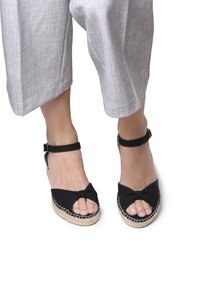 Must have espadrilli sandaalid musta värvi, millel on paeltega detail, pahkluu rihm ja jute-sisuga tald. Kannab heledat halli, laia jala lõikega pükse.
