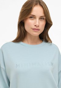 Jauna moteris su šviesiai rudais plaukais, vilkinti šviesiai mėlyną džemperį su užrašytu "MINIMALIZMAS" ant krūtinės.