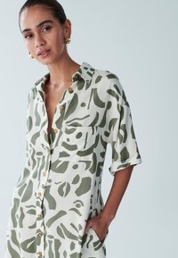 Sukienka koszulowa z lekkiego materiału, z zielonym abstrakcyjnym wzorem na jasnym tle, z krótkimi rękawami i zapięciem na guziki z przodu.