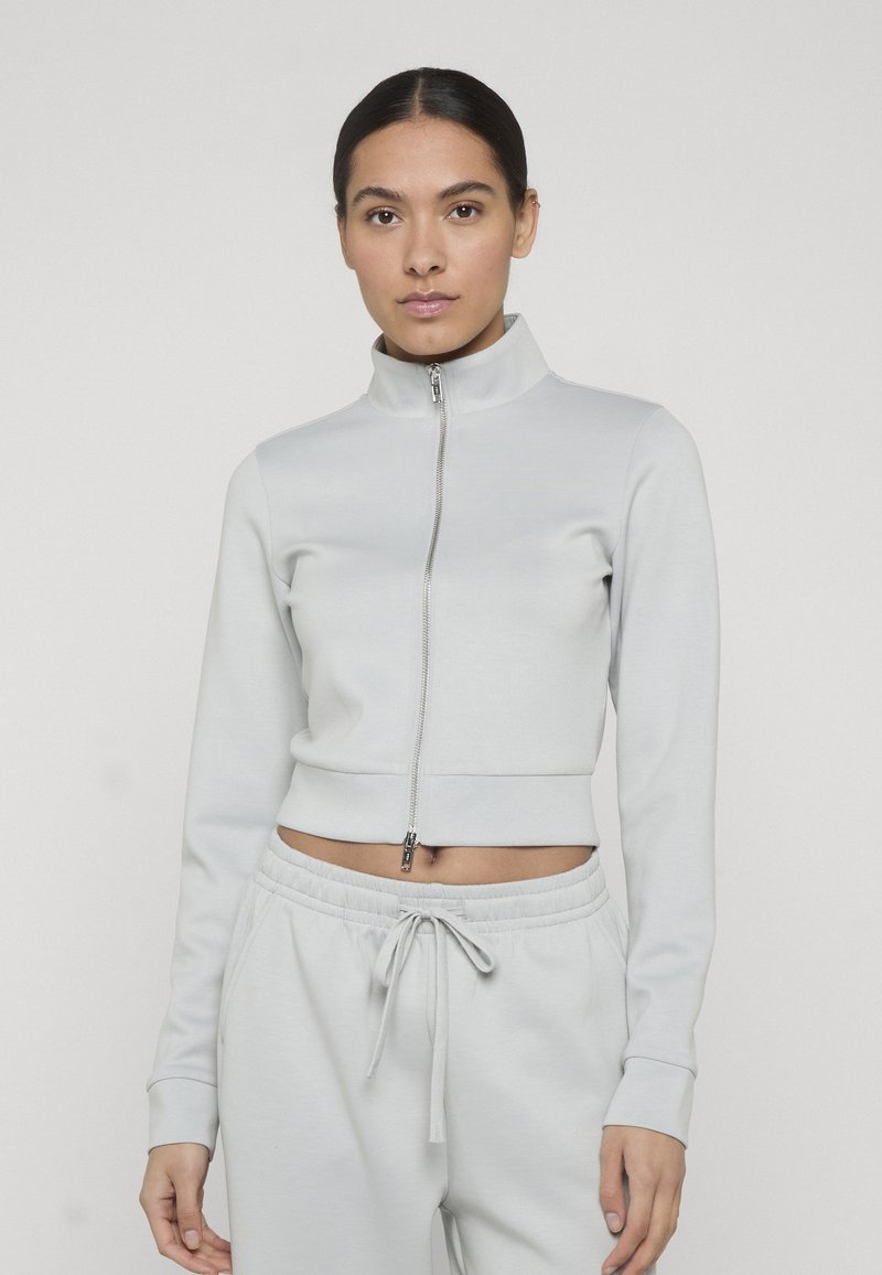 Jeune femme portant une veste courte gris clair à fermeture éclair et un pantalon de jogging assorti avec cordon de serrage, sur un fond uni.