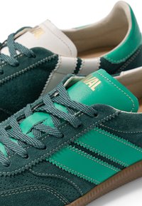 Baskets en daim vert et blanc avec des bandes latérales en zigzag et des lacets verts, partiellement superposées sur un fond blanc.