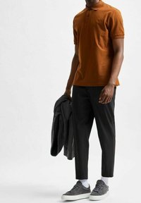 Brun polo-tröja med två knappar, matchad med svarta smalrandiga byxor. Grå sneakers och en grå jacka draperad över en arm.