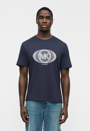Mann trägt ein marineblaues Michael Kors T-Shirt mit weiß gestreiftem, runden Logo und hellblauen Jeans, steht vor einem einfarbigen Hintergrund.