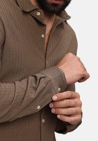 Camicia button-up marrone con texture, colletto e maniche lunghe. Presenta un tessuto a costine e bottoni di colore chiaro, mostrando una vestibilità rilassata.