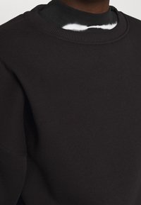 Sudadera negra con un cuello redondo acanalado, de textura suave y corte relajado. Superpuesta sobre una camiseta negra con un patrón de tinte anudado.