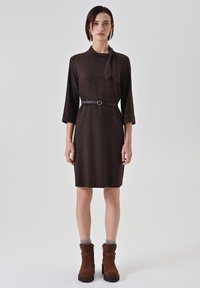 Robe marron longueur genou avec un détail de lien au niveau du col, manches trois-quarts et ceinture contrastante ; associée à des bottines marron.