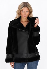 Donna che indossa un giubbotto in similpelle nera e pelliccia, tiene il colletto con una mano, vestita con jeans blu su sfondo bianco.