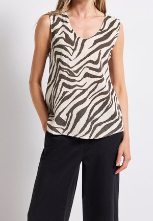 Bez rękawów top w zebrowy wzór w połączeniu z luźnymi czarnymi spodniami, noszony przez stojącą osobę z jedną ręką w kieszeni na jednolitym tle.