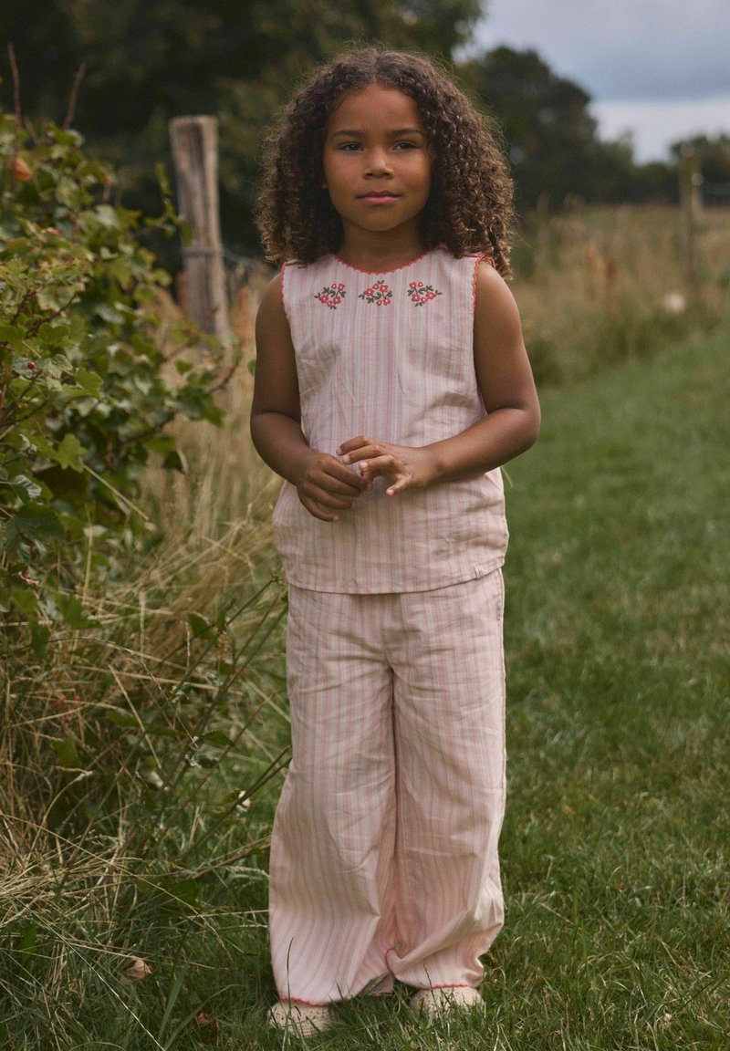 Jeune fille aux cheveux bouclés debout sur un chemin herbeux, portant un top sans manches rose pâle rayé et un pantalon large avec broderie florale.