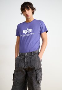 Purple cotton t-shirt with a white logo featuring an 'A' and three stripes, paired with dark grey cargo pants with multiple pockets.