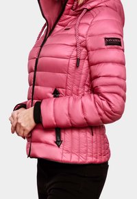 Rosa quilted jakke med hætte og sorte lynlåsdetaljer, båret af en person med hænderne samlet nær livet og sorte bukser synlige.