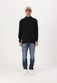 Fekete pulóver magas, zippzárral ellátott gallérral, kék farmernadrággal és fehér sneakers-rel párosítva. A modellen világosszürke sapka van.